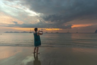 Woman, 60-65, taking a photo of the sunset with her smartphone, Koh Mook Island, Andaman Sea,