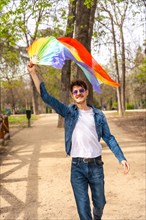 Vertical photo of a gay man raising and waving a rainbow colored hand fan in a park