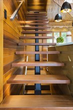 Looking up illuminated wooden staircase with opened steps and clear glass railing leading to