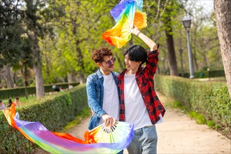 Multiracial gay couple waving lgbt rainbow fans while walking together along a public park