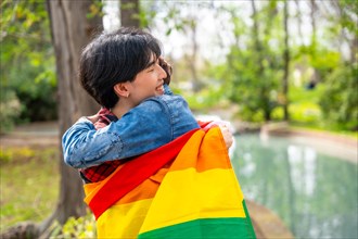 Multiracial gay couple embracing inside a lgbt flag standing in a park