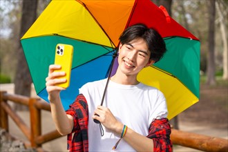 Chinese gay man taking a selfie with mobile phone while holding a rainbow colored umbrella standing