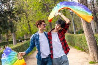Multiracial gay couple celebrating love waving lgbt rainbow fans in a park