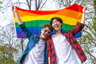 Low angle view portrait of a happy gay couple laughing while raising lgbt rainbow flag outdoors in