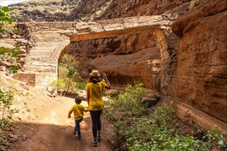 A woman and a child are walking through a rocky area. The woman is wearing a yellow shirt and a hat