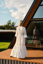 Barefoot woman standing on wooden terrace of tiny house and looking at her reflection in large