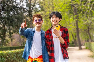 Multi-ethnic smiling gay couple standing pointing ahead using phone in a park