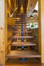 Looking up illuminated wooden staircase with opened steps and clear glass railing leading to