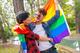 Happy and romantic multi-ethnic gay couple embracing wrapped in rainbow flag in a park
