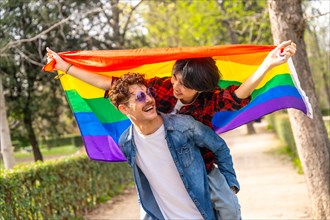 Multi-ethnic gay couple piggybacking in a park raising a lgbt rainbow flag