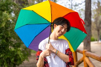 Cute gay chinese man smiling at camera holding a rainbow colored umbrella in a park
