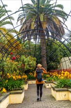 A woman wearing a hat and a backpack walks through a garden. The garden is filled with yellow