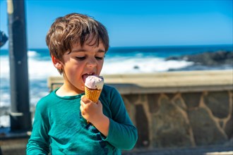 A young boy is eating an ice cream cone on a beach. The boy is smiling and enjoying his treat