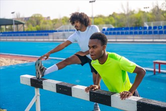 Two young african american runners stretching legs in a fence in a running track