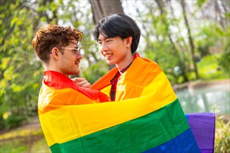 Multi-ethnic gay couple embraced in a park wrapped in rainbow flag