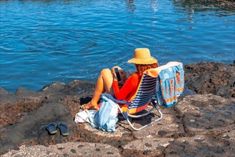A woman is sitting in a chair by the water, wearing a straw hat and holding a cell phone