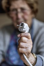 Senior citizen holding a power cable with plug in her hand at home, symbolising energy costs and