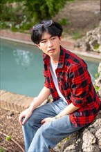 Vertical photo of an Asian gay man sitting next to urban stream of water in a park