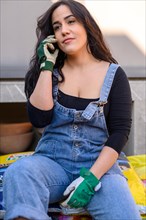 A woman in denim overalls and gloves sits casually outdoors with a relaxed posture