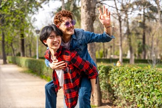 Horizontal photo with copy space of a gay couple having fun in a park waving and piggybacking