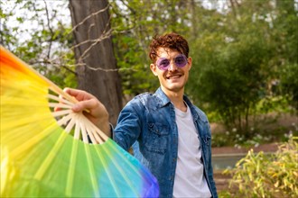 Gay man in sunglasses holding a rainbow fan in a park