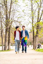 Vertical full length photo of a gay male couple walking along a park carrying LGBT rainbow flag