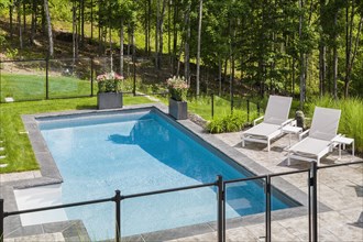 Two white long chairs on edge of in-ground swimming pool enclosed by clear glass and black metal