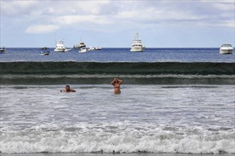 San Juan del Sur, Nicaragua, swimmers in the water in front of a big wave, boats and blue sky in