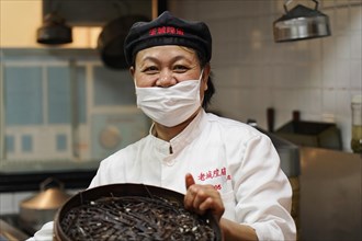 Shanghai, China, Asia, A smiling cook with a mask stands in a kitchen and holds a pan with