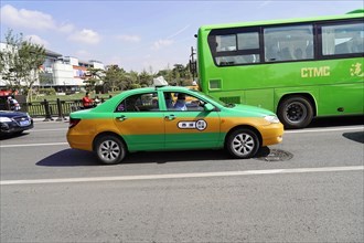 An orange taxi and a green bus drive along a city street