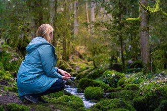 A woman sits on the bank of a forest stream, surrounded by moss and green vegetation, and seems to