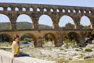 Woman sitting on a wall in front of the Pont du Gard, Roman aqueduct over the river Gardon,