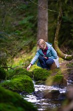 A woman carefully crosses a small, moss-covered stream in the forest, Calw, Black Forest, Germany,