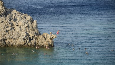 People jumping from cliffs into the sea, surrounded by swimmers in clear water, Lindos, Rhodes,
