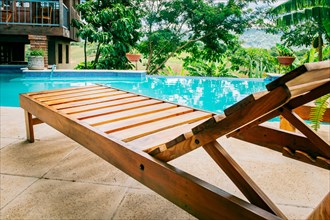 A wooden deck chair near a swimming pool. View of a Deck chair near a beautiful pool surrounded by