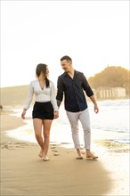 Vertical photo of caucasian young romantic couple holding hands walking on a beach enjoying sunset