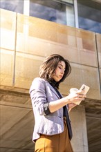 Young business woman walking down the street holding cell phone and glass of coffee, A Woman