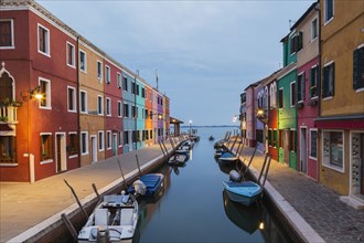 Moored boats on canal lined with colourful stucco houses and shops at dusk, Burano Island, Venetian
