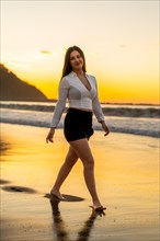 Vertical portrait of a chic sensual woman walking barefoot along the beach during sunset