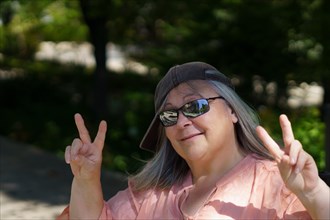 Cheerful and funny older gray-haired woman with cap and sunglasses making a victory sign