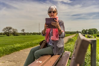 Senior citizen sitting on a bench and looking at her smartphone, frontal view, Deichkrone, Rhine,