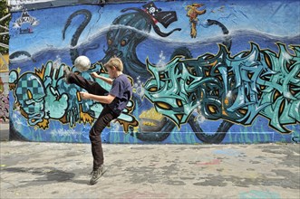 A ten-year-old boy plays with his football in front of a graffiti wall, Germany, Europe