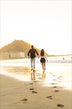 Vertical rear view of a tender young couple walking holding hands on the edge of the sea water