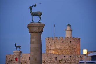 Bronze deer on pedestal in the foreground with old fortress walls and lighthouse in the background,