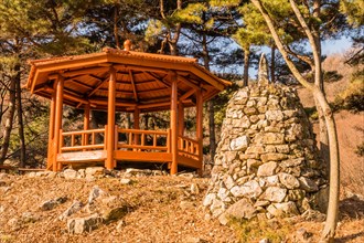Stone pyramid next to wooden gazebo on small hill under grove of evergreen trees located in public