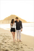 Vertical photo of a romantic Young couple walking embraced barefoot along a beach during sunset