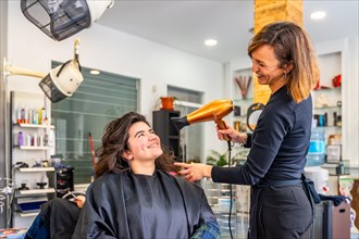 Complicity between smiling hairdresser and female customer sitting while drying the hair in the