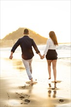 Vertical photo of the rear view of a playful couple running holding hands on the edge of the sea