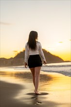 Vertical rear view of a sensual elegant woman walking barefoot along the beach during sunset
