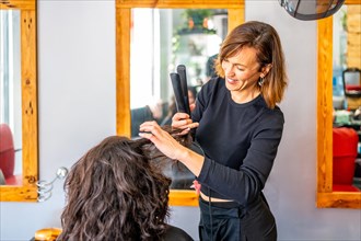 Happy hairstylist attending a female client sitting next to a mirror ironing the hair